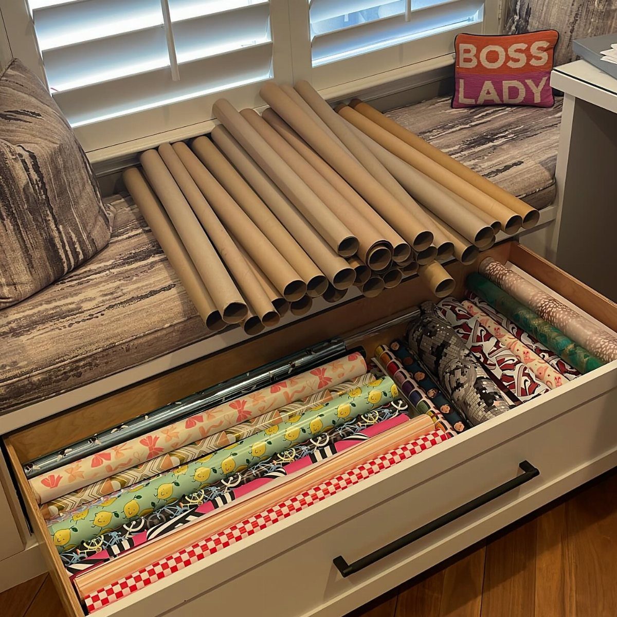 Colorful wrapping paper rolls organized inside a white wooden drawer under a window seat.