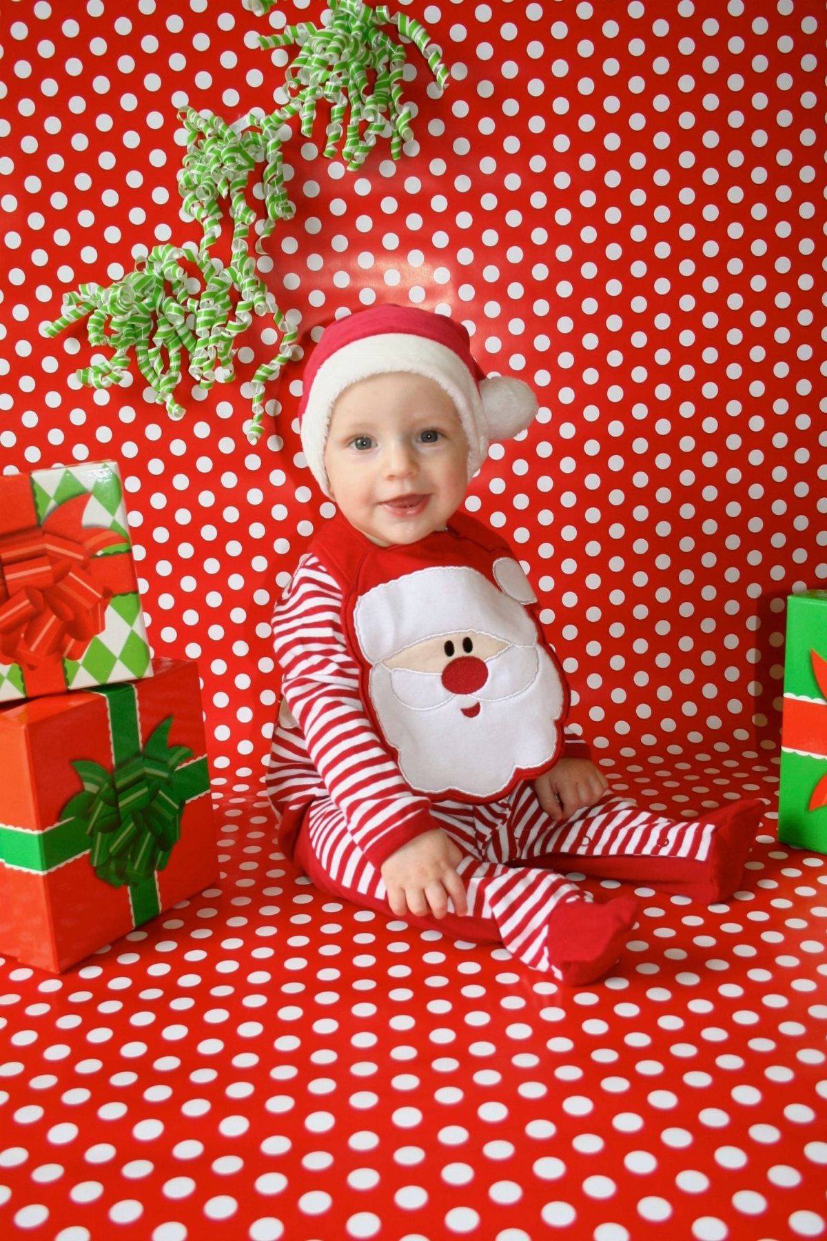 Baby in Santa outfit sitting against a red polka-dot wrapping paper backdrop
