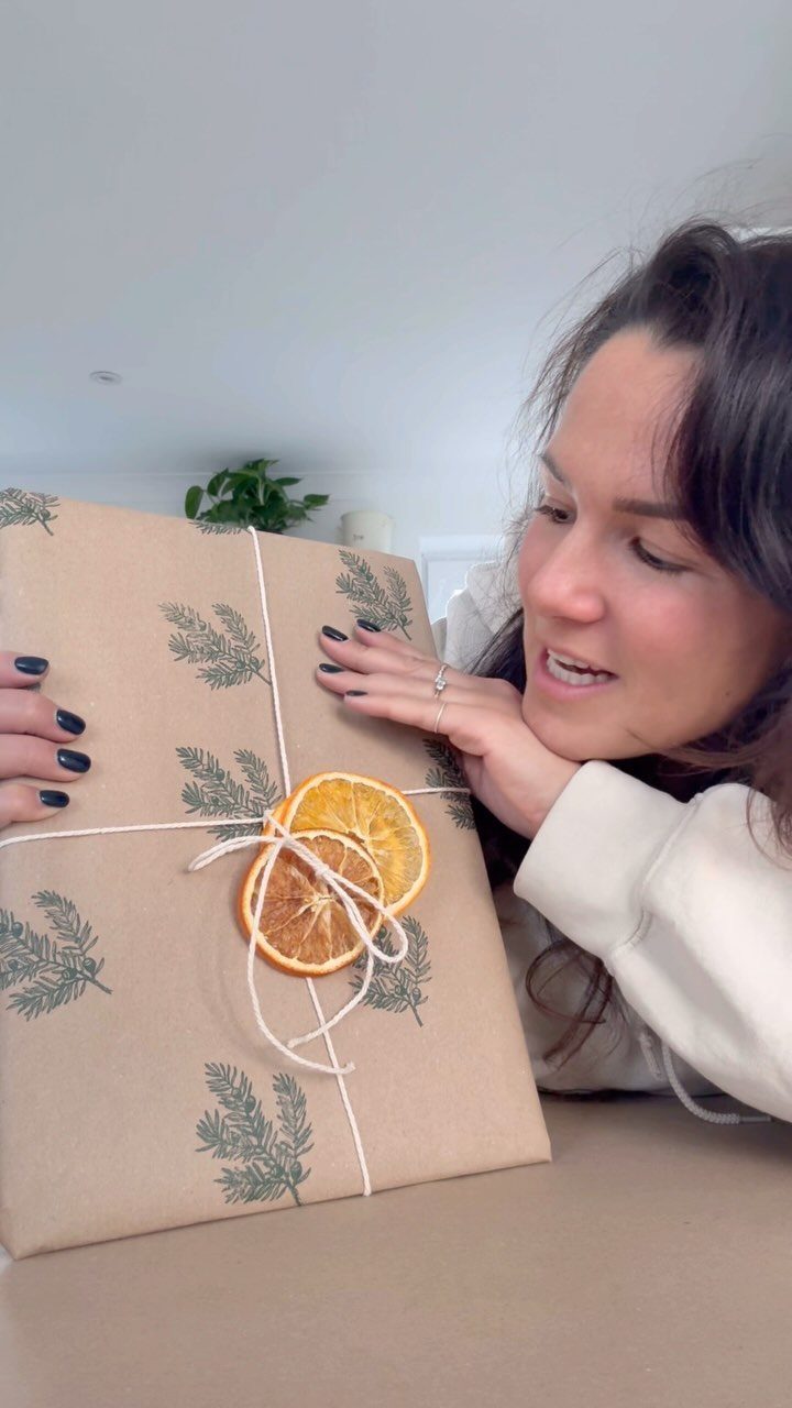 Woman admiring a kraft paper wrapped gift decorated with pine prints and dried oranges.